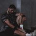 A muscular man in a gym environment taking a moment of rest, reflecting in a dark moody setting.